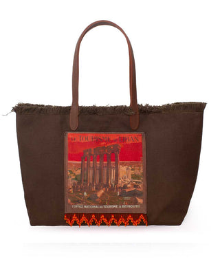 A dark brown canvas tote bag with frayed edges, brown leather handles, and a central graphic print depicting the Baalbeck in lebanon under a red sky. The bottom edge features a red and black geometric patterned fringe.