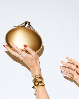 Gold handbag with two silver bird on top held by a hand with red nail polish against a white background.