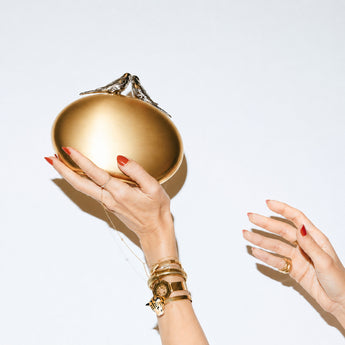 Gold handbag with two silver bird on top held by a hand with red nail polish against a white background.