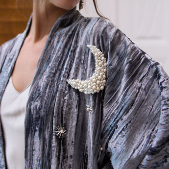 Close-up of a person wearing a blue gray velvet jacket with decorative brooches, including a moon-shaped pearl brooch.
